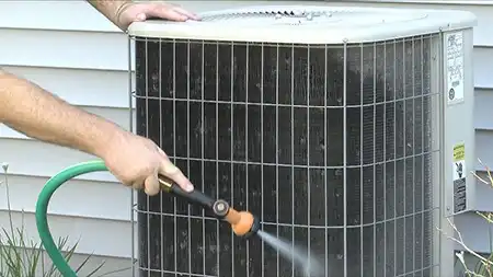 Person gently spraying water from a hose on a dirty outdoor AC condenser coil to clean the fins.